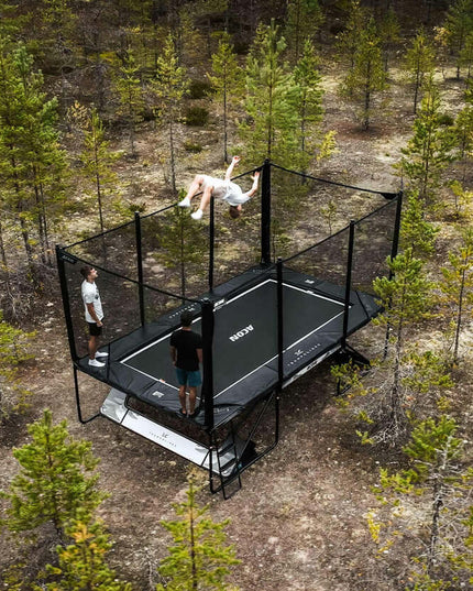 Luftaufnahme eines ACON X Trampolins mit Sicherheitsnetz im Kiefernwald, auf dem mehrere Personen springen und trainieren.