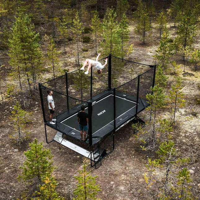 Luftaufnahme eines ACON X Trampolins mit Sicherheitsnetz im Kiefernwald, auf dem mehrere Personen springen und trainieren.