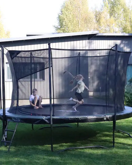 Zwei Kinder nutzen ein ACON AIR Gen 2.0 Rundtrampolin mit Sicherheitsnetz auf einer Rasenfläche im Garten.