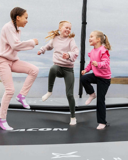Drei Kinder springen und spielen auf dem schwarzen ACON X Trampolin mit Sicherheitsnetz im Freien.