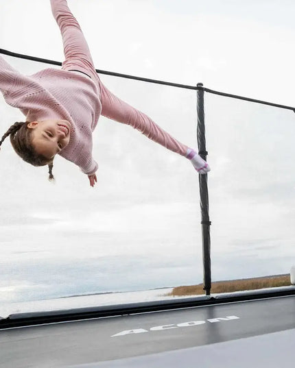 Kind springt kopfüber auf dem ACON X Trampolin mit Sicherheitsnetz, während zwei weitere Kinder zusehen.