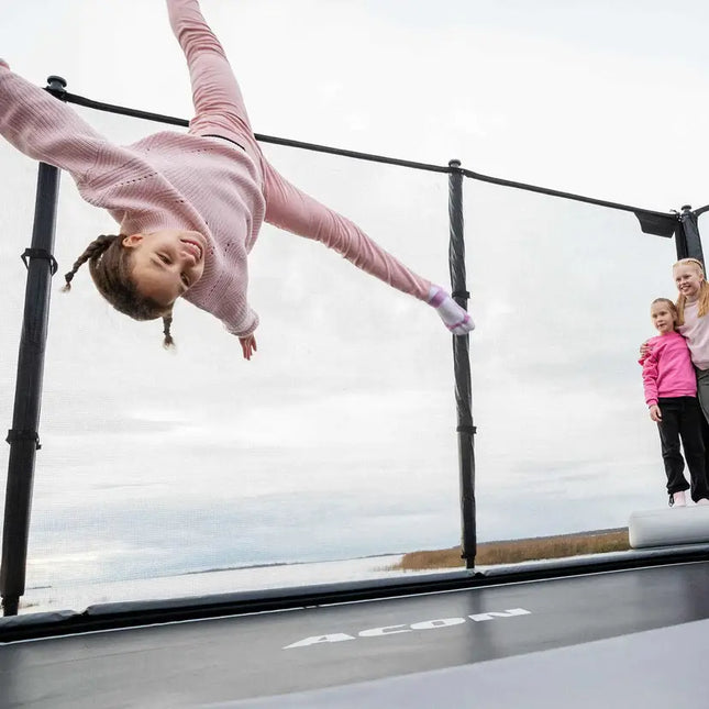 Kind springt kopfüber auf dem ACON X Trampolin mit Sicherheitsnetz, während zwei weitere Kinder zusehen.