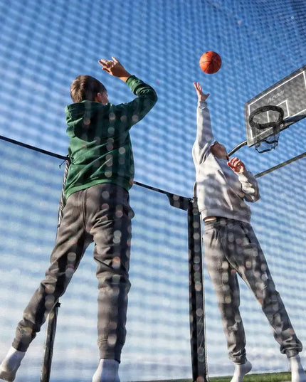 Zwei Kinder spielen Basketball auf einem ACON X Trampolin mit Sicherheitsnetz und befestigtem Basketballkorb.
