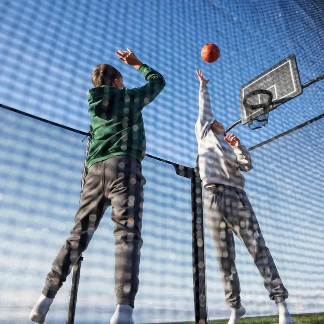 Zwei Kinder spielen Basketball auf einem ACON X Trampolin mit Sicherheitsnetz und befestigtem Basketballkorb.
