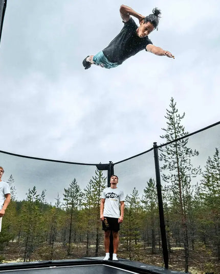 Athlet führt einen hohen Sprung über einem ACON X Trampolin mit Sicherheitsnetz aus, während zwei Personen auf der Sprungfläche zusehen.