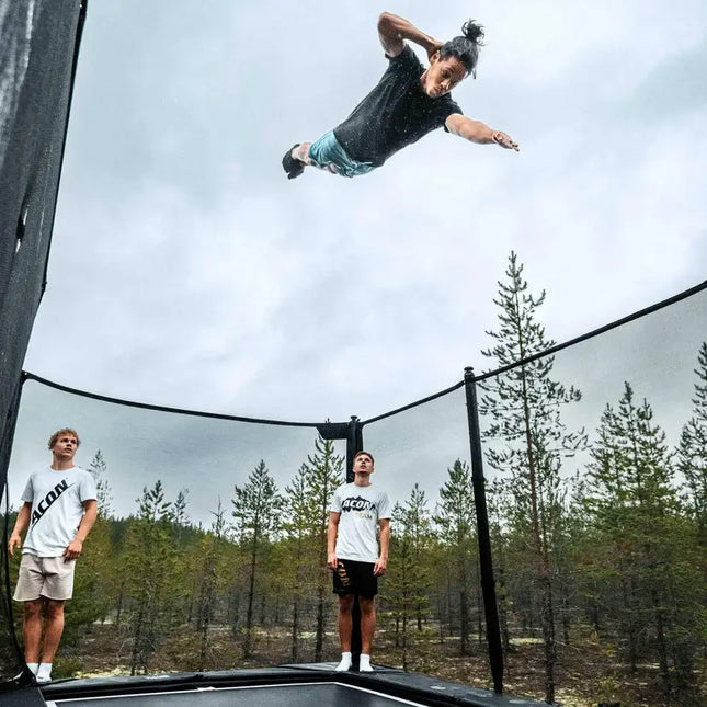 Athlet führt einen hohen Sprung über einem ACON X Trampolin mit Sicherheitsnetz aus, während zwei Personen auf der Sprungfläche zusehen.